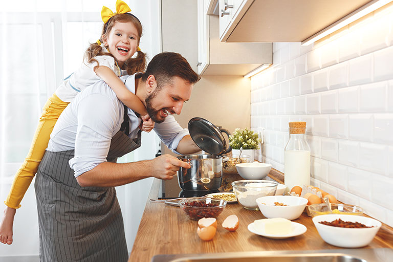 Man preparing dinner in a pot on the stove with a young girl hanging on his back