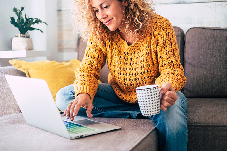 Woman with long curly hair sitting on couch while holding a coffee mug and using a laptop