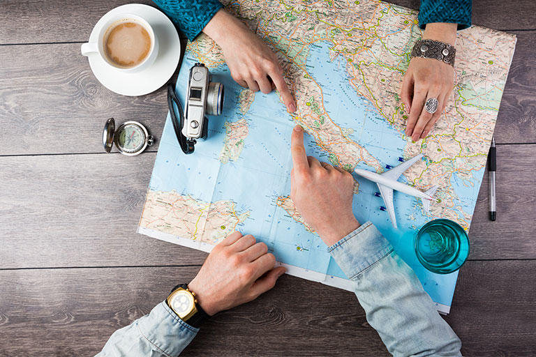 Map of the world laying on a dark wood table with coffee, camera, compass, and two people's hands pointing at a destination
