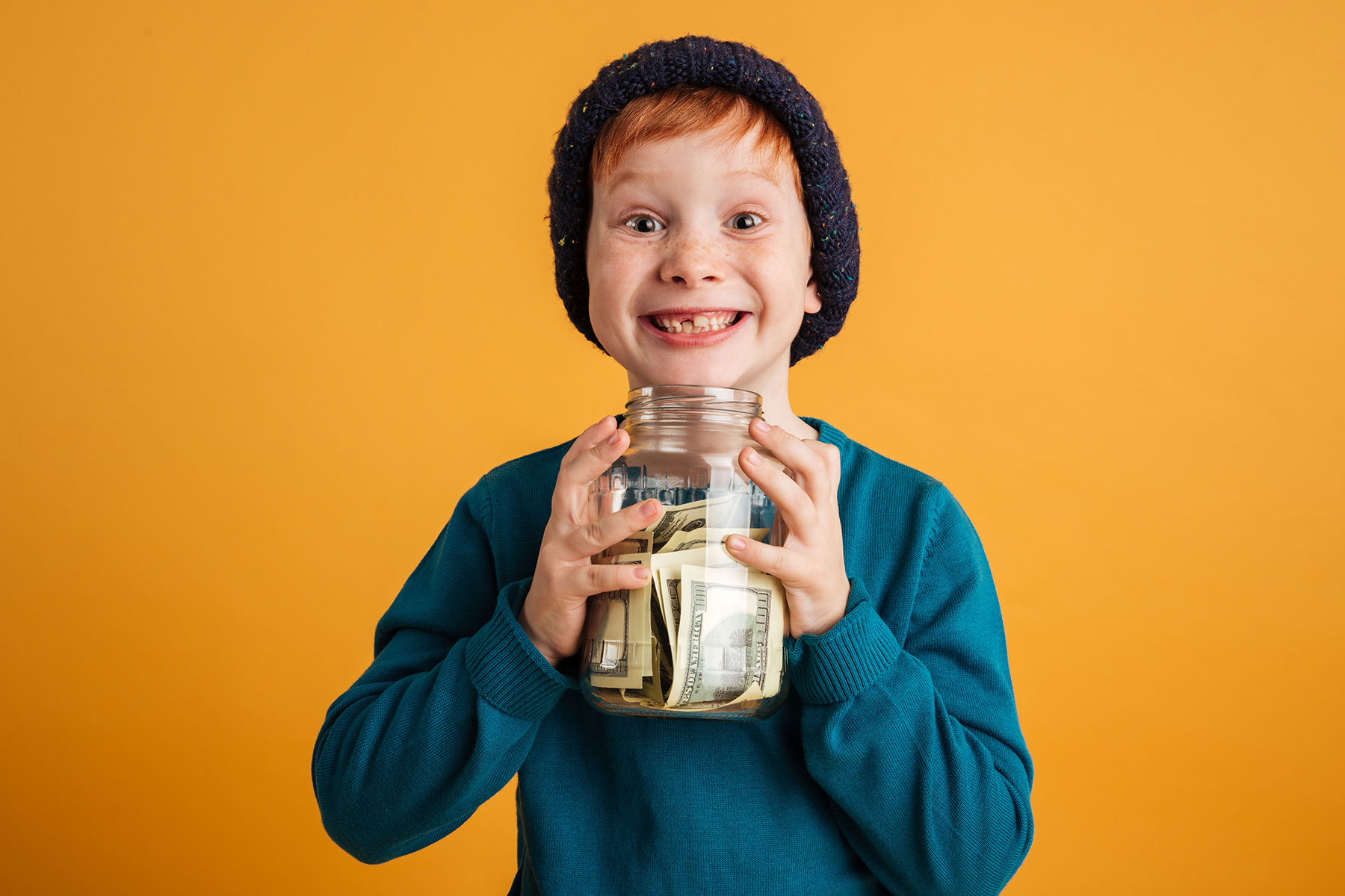 Young boy with red hair and a beanie on holding a large clear jar  filled with cash in front of a yellow background