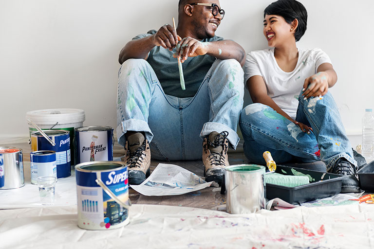 Man and woman sitting in the floor of home holding a paintbrush with buckets of paint around them