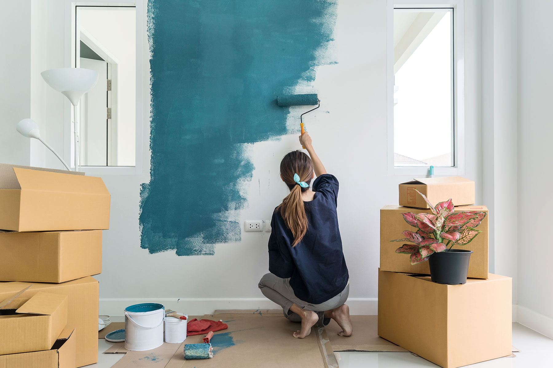 Young woman with hair in ponytail painting interior wall teal with paint roller in house