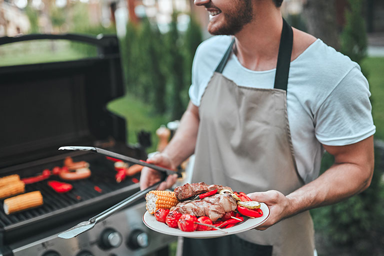 Bearded man in apron standing outside on patio near grill holding a plate of meat and vegetables and tongs