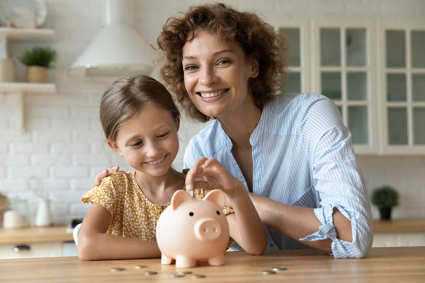 Happy smiling mom cuddling young smiling girl putting coin into pink piggybank.