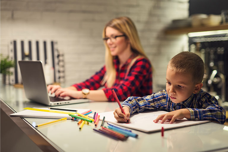 Young boy using colored pencils to color a picture in the kitchen with mom working on a laptop next to him