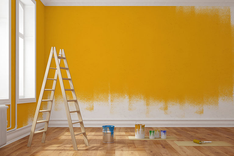 Partially painted yellow wall in home with ladder and buckets of paint