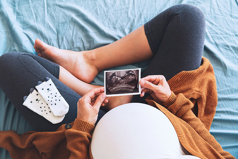 Pregnant woman sitting with crossed legs holding picture from ultrasound with baby socks on her leg